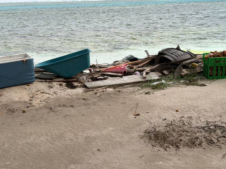 Trash and broken items scattered on a sandy shoreline beside greenish sea water