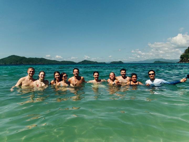 Group of smiling travelers waist-deep in clear tropical sea with green islands on the horizon.
