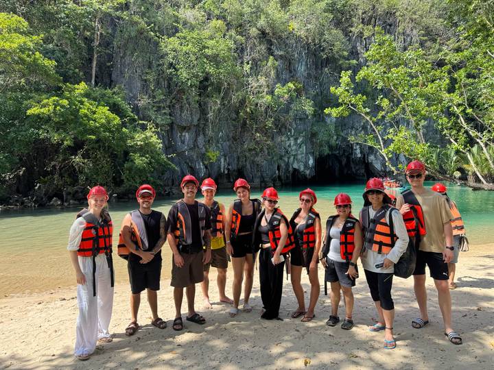 A tour group wearing red helmets and life-jackets stands on a sandy shore in front of a turquoise river cave surrounded by lush jungle cliffs.