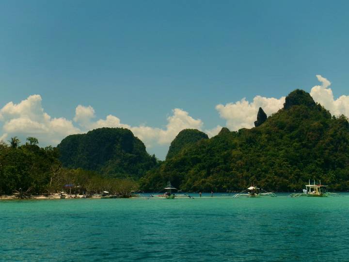 Turquoise sea with small outriggers anchored by lush jungle-clad limestone islands under a clear sky.