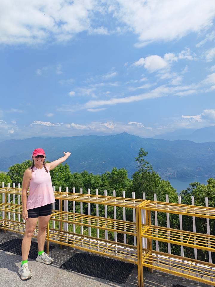 A smiling woman in a pink top and cap points toward snow-capped Himalayan peaks above a green valley and lake.