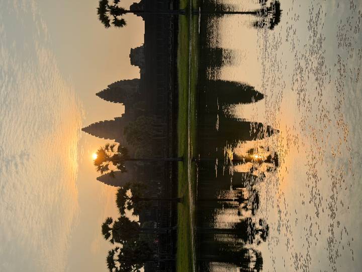 Angkor Wat’s iconic towers silhouette against a pastel sunrise, their reflection shimmering in the still lotus pond.