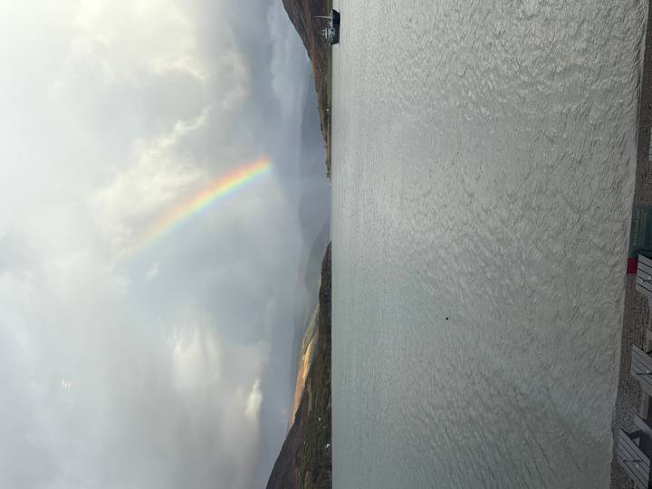 Soft rainbow arcs over a tranquil Scottish loch framed by misty hills and grey clouds.