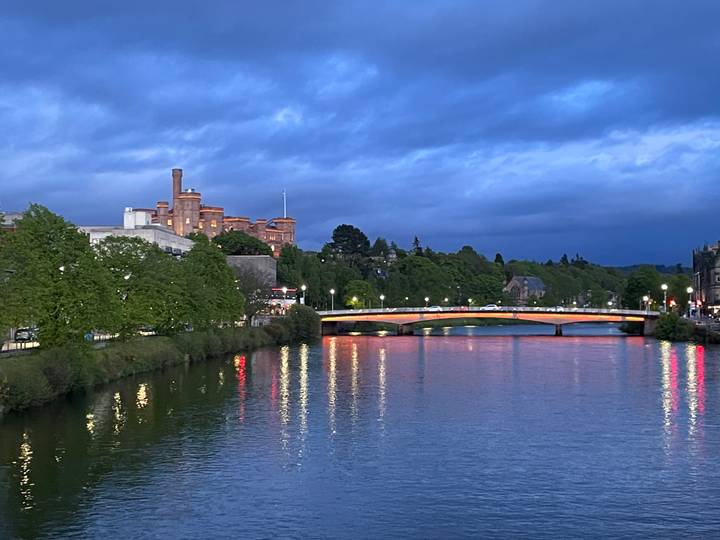 Evening lights reflect in the River Ness below a bridge with Inverness Castle silhouetted against a deep blue twilight sky.