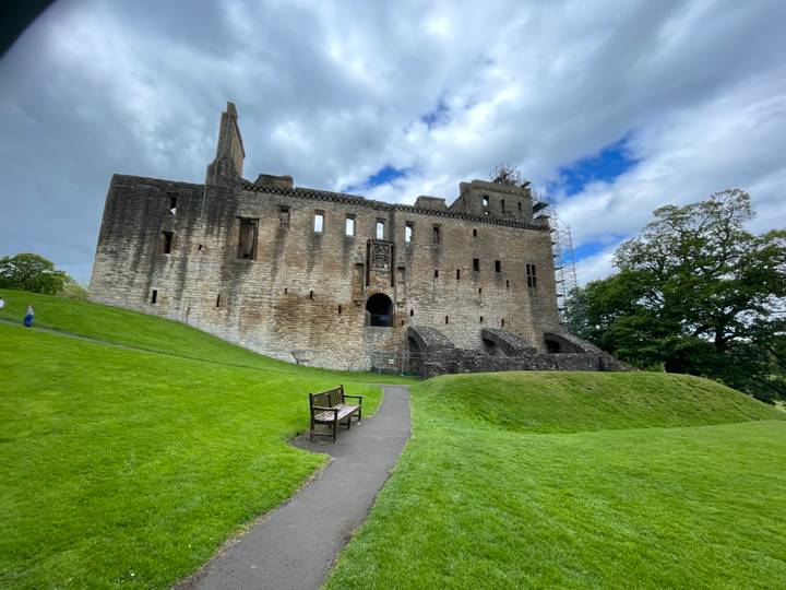 The weathered stone walls of Linlithgow Palace stand on a grassy hill under a partly cloudy sky, with a lone visitor nearby.