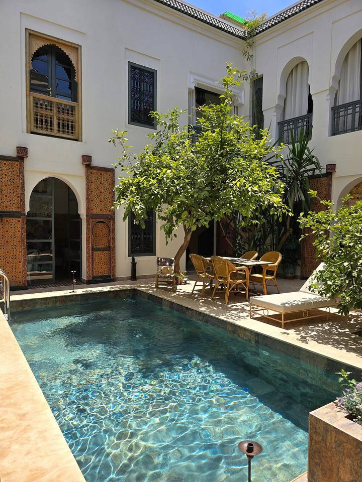 Tranquil riad courtyard with mosaic walls, a small pool, citrus tree shade, wicker chairs and a sun lounger in Marrakesh.