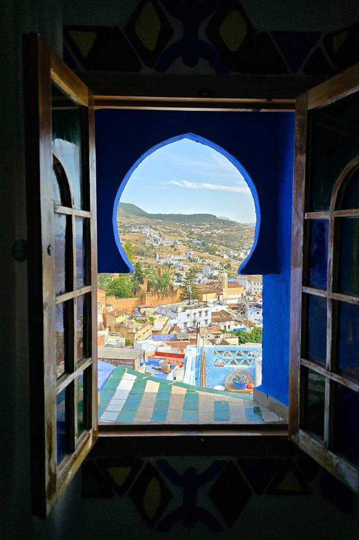 View through a Moorish horseshoe-shaped window to a hillside town of white and blue houses under a clear sky.