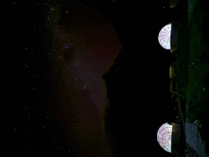 Star-filled Andean night sky over dark mountain silhouette with two illuminated geodesic dome tents below.