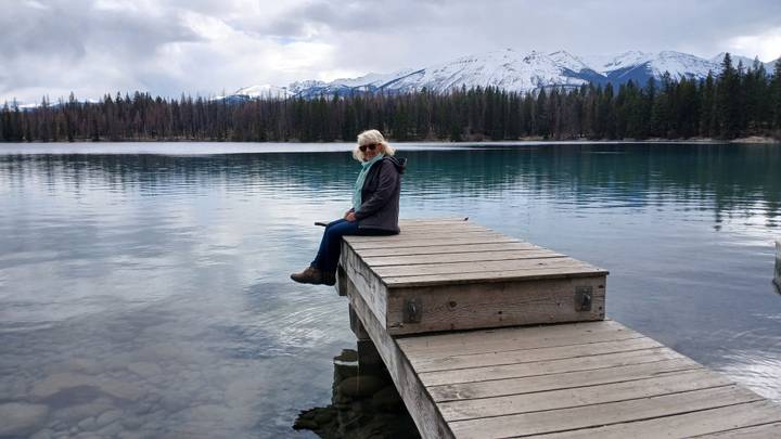 Woman sits quietly on a wooden dock extending into a glassy alpine lake surrounded by forested peaks.