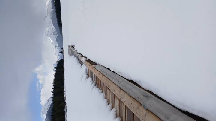 Narrow wooden boardwalk disappearing into snow-covered lake plain with distant alpine peaks beneath mixed clouds.