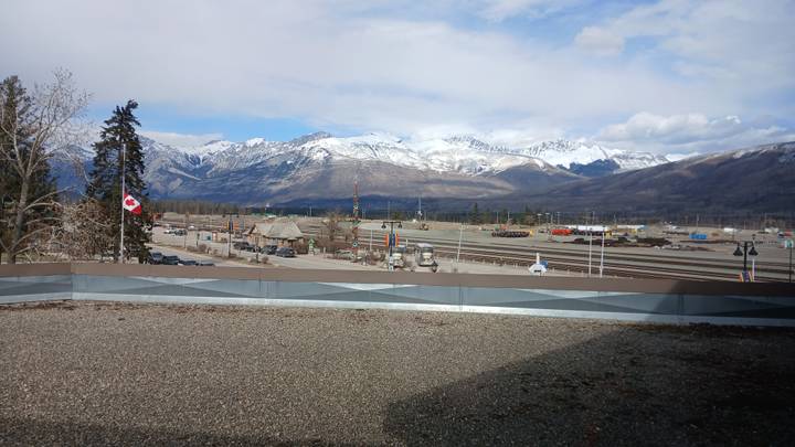 Rail yard and Canadian flag with a backdrop of snow-clad Rockies under partly cloudy sky.