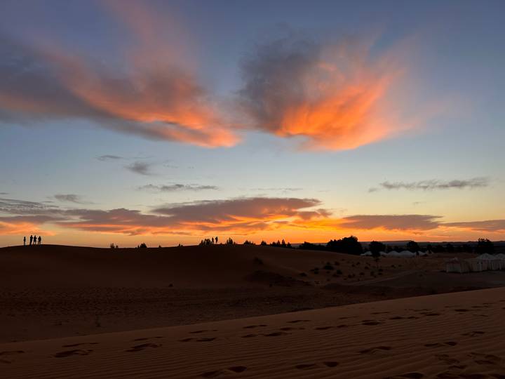 Fiery sunset clouds over golden Saharan dunes with silhouettes of travelers on the ridge.
