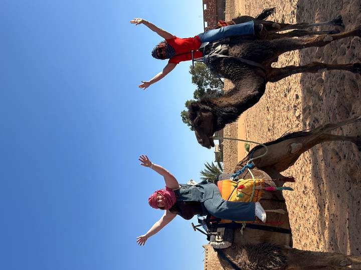 Two excited riders raise their arms atop camels against a deep blue desert sky.