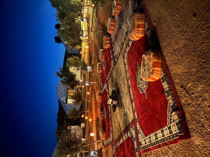 Atmospheric desert camp at night with carpets, fire pit and lanterns glowing under deep blue sky.
