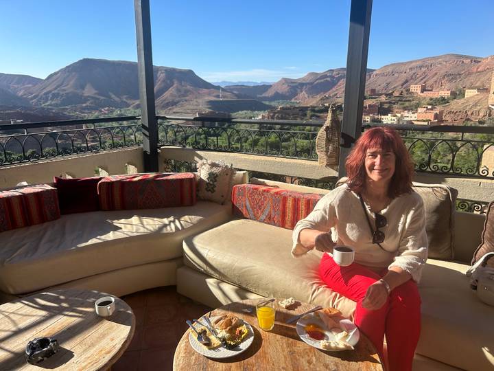 Woman enjoys breakfast coffee on a cushioned terrace overlooking rugged red mountains and a green valley.