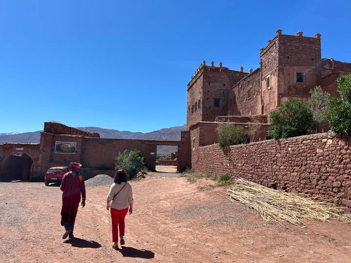 Visitors follow a local guide toward the earthen walls of Ait Benhaddou kasbah under blue sky.