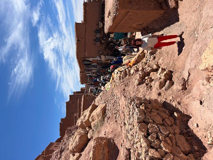 Market alley climbing the red-stone slope of Ait Benhaddou with craft stalls and blue sky.