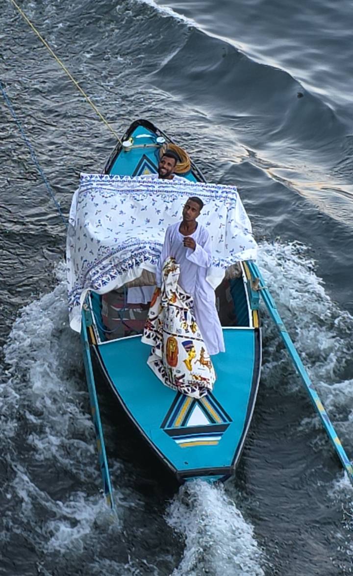 Man in traditional white robe standing on the bow of a small boat decorated with patterned cloth on the Nile