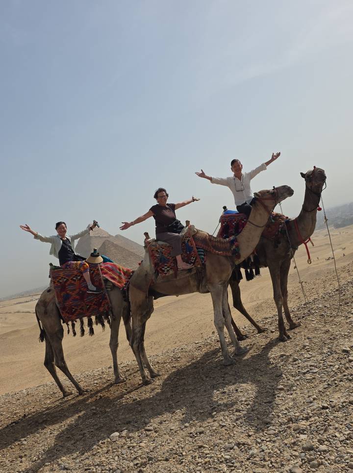 Three travellers joyfully pose on camels with the Great Pyramid rising in the desert background