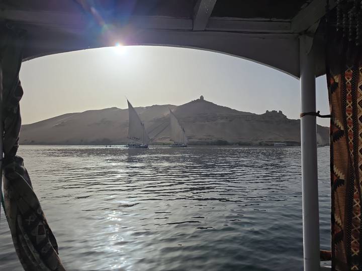 View from a felucca of two sailboats gliding across the Nile with a sand dune and tomb in the distance