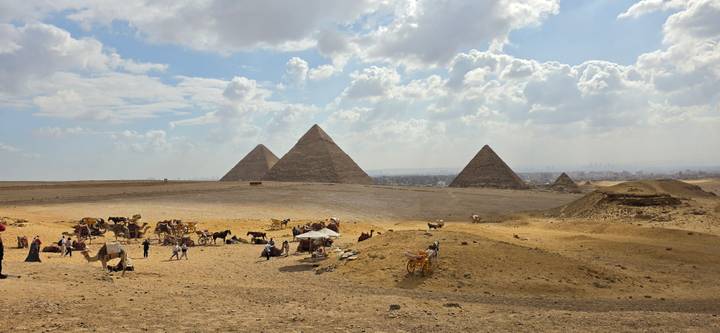 The iconic pyramids of Giza rise above a sandy plateau dotted with camel caravans under a dramatic sky.