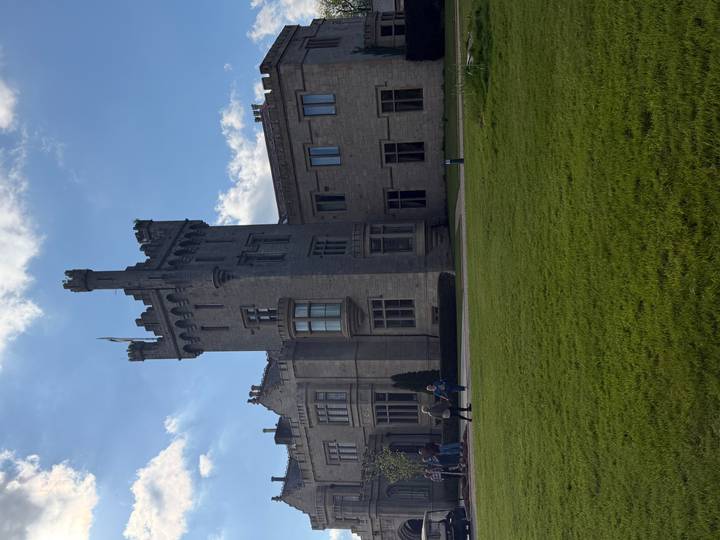 A grey-stone baronial castle with a central tower and manicured lawn under a bright blue sky with passing clouds.