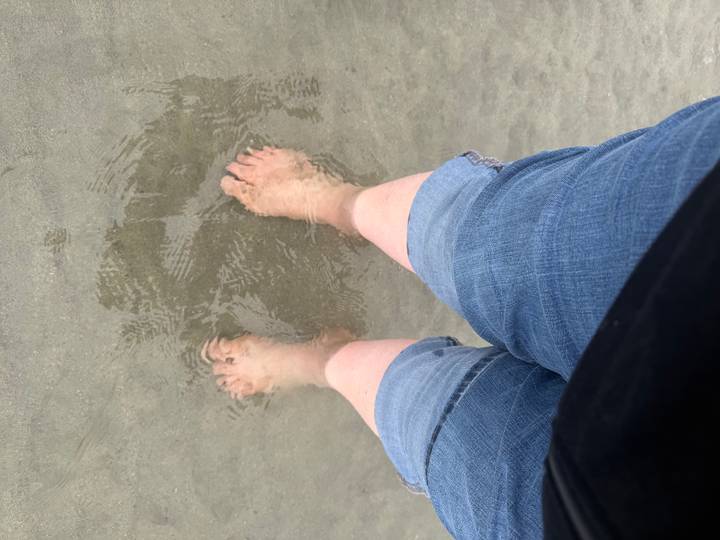 Bare feet in rolled-up jeans standing in shallow, rippling seawater on a sandy beach.