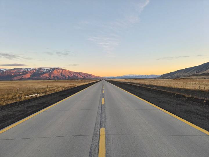 A long straight highway disappears into a pastel sunrise horizon between rugged Patagonian ranges.