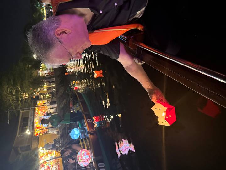 At night on Hoi An’s river, a man releases a colourful paper lantern from a wooden boat amid glowing lights.