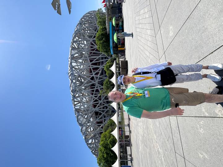 Smiling couple posing in front of Beijing’s Bird’s Nest Olympic Stadium under a bright blue sky.