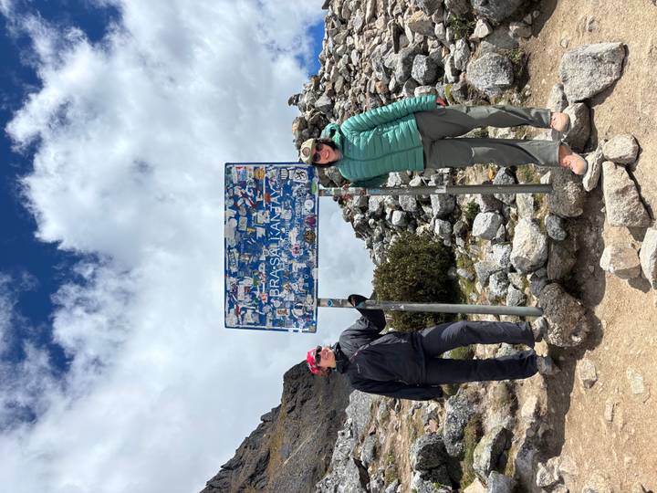 Two hikers pose at the Salkantay Pass sign surrounded by rocky scree and cloudy skies