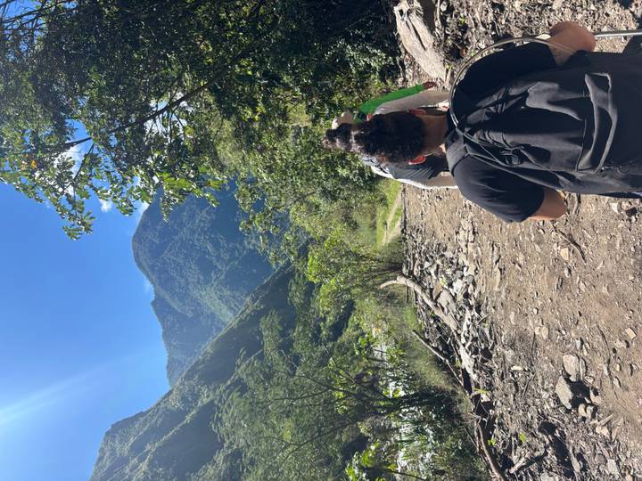 Trekkers follow a rocky jungle path with steep green mountains rising ahead in bright sunshine