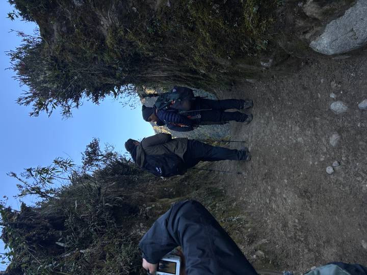 Two trekkers pause between high earthen walls on a narrow mountain trail in cool morning light