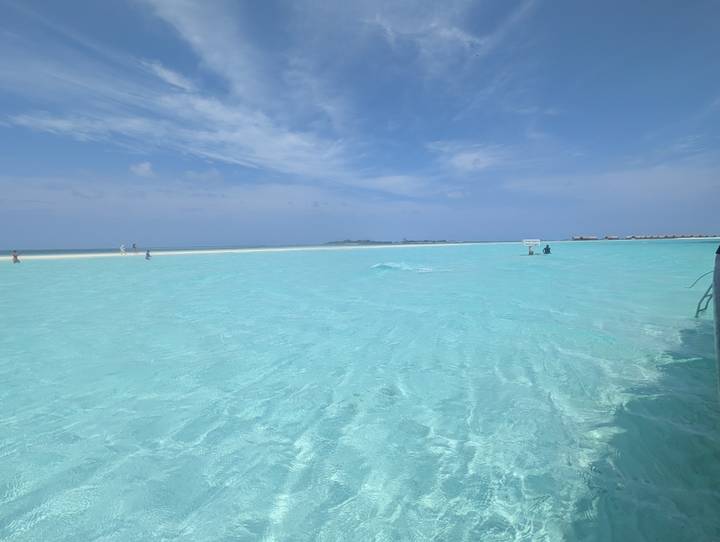 Crystal-clear turquoise lagoon water stretches toward a distant sandbar and tiny resort huts in the Maldives.