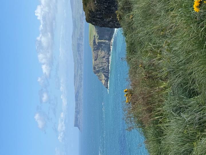 Dramatic sea cliffs and turquoise waters stretch beneath a blue sky dotted with white clouds.