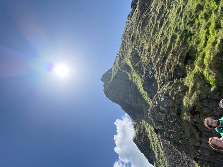 A steep grassy peak rises toward the bright noon sun with a few hikers at its rocky base.