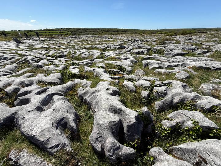 The stark limestone pavement of the Burren stretches into the distance under a bright sky with tiny hikers.