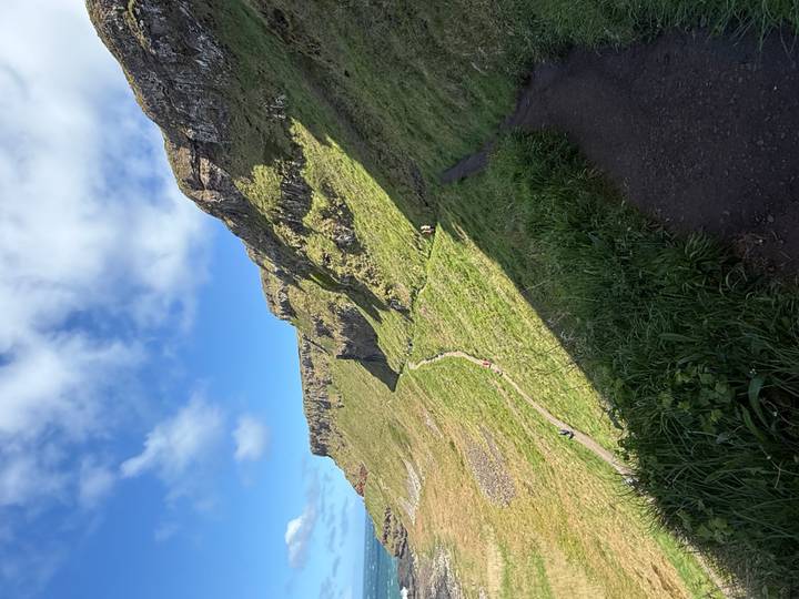 A winding cliffside hiking trail leads visitors through green slopes under a bright sky.