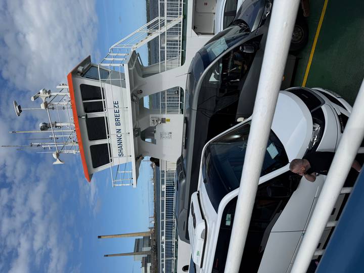 Vehicles and a passenger stand on the 'Shannon Breeze' ferry deck beneath communication masts.