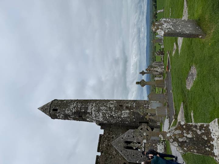 Ancient round tower and Celtic crosses dominate a grey-stone graveyard under a cloudy sky.