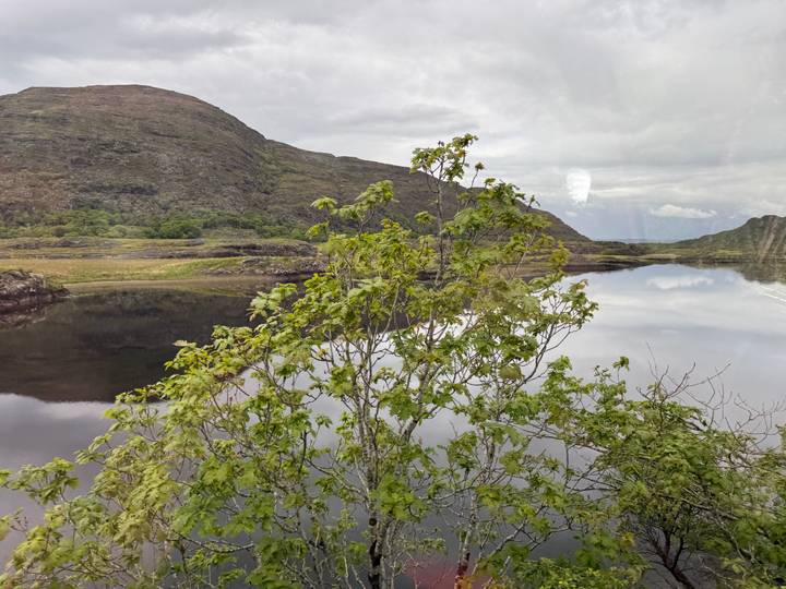 A reflective lake and rugged hills are seen through a bus window, faint reflections visible on the glass.