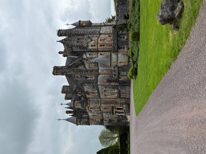 Large stone Victorian-style castle with turrets and chimneys set beside a manicured green lawn under an overcast sky.