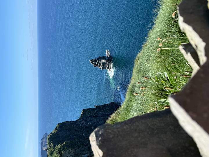 Dramatic ocean cliff with a single sea stack rising from deep blue Atlantic waters on a clear sunny day.