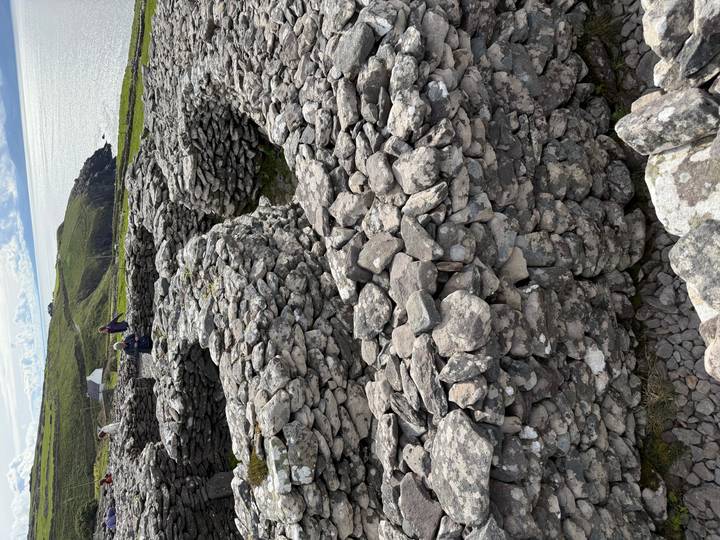 Ancient circular stone huts and walls overlooking green coastal hills with visitors exploring the archaeological site.