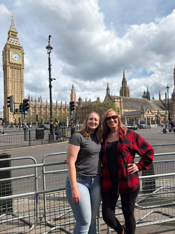 Two travellers smile in front of London’s Houses of Parliament and Big Ben on a partly cloudy day.