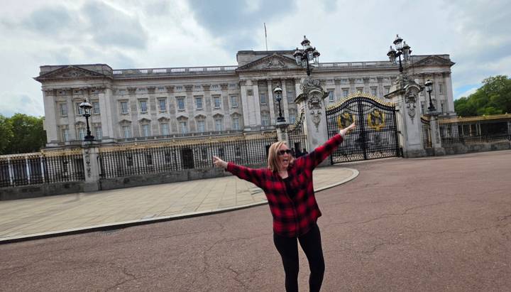Happy traveller stretching arms before Buckingham Palace’s ornate gates and façade under dramatic clouds.
