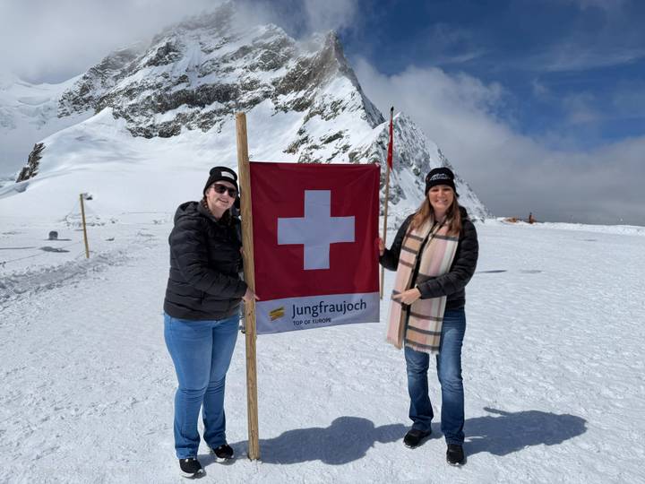 Two women holding a Swiss flag on the snowy plateau of Jungfraujoch with alpine peaks behind.
