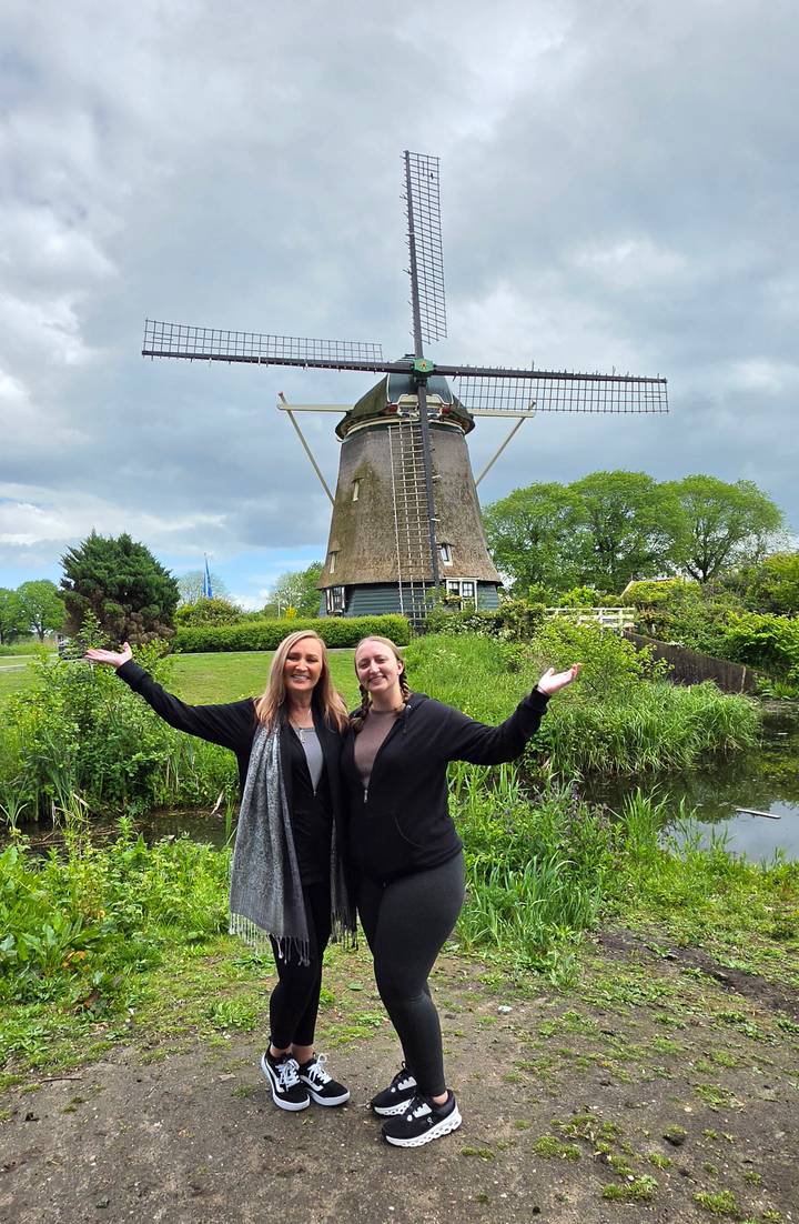 Travellers stand with arms outstretched in front of a traditional Dutch windmill surrounded by lush greenery.
