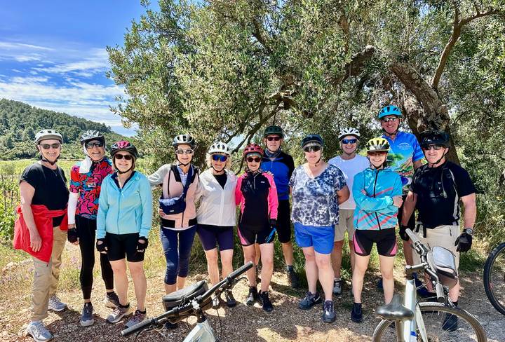 A smiling cycling group in helmets poses under an olive tree with bikes.