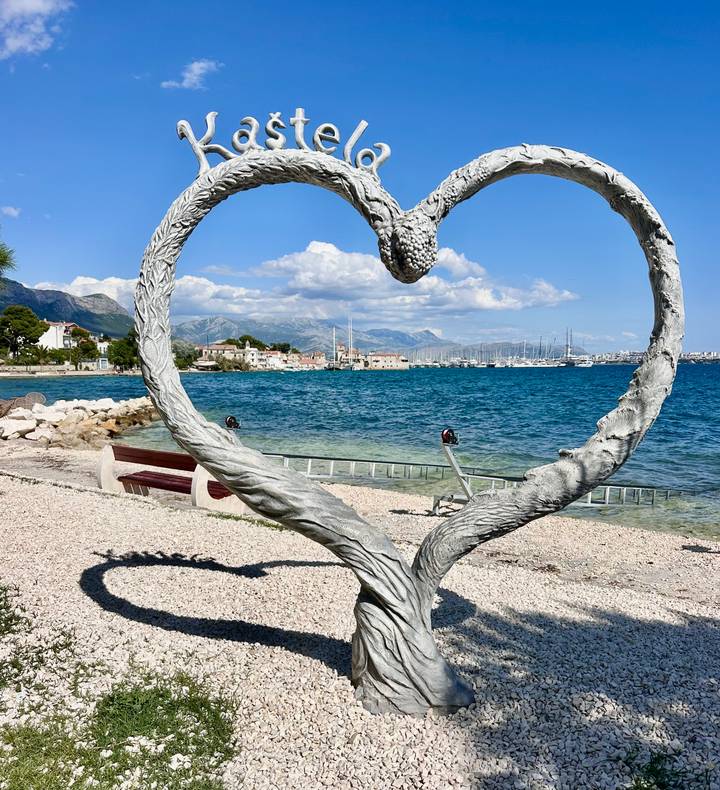 A heart-shaped metal sculpture frames a coastal Croatian town and marina across turquoise water.